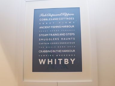 A framed blue poster with white text listing Whitby attractions and features at West Cliff Apartment in Whitby