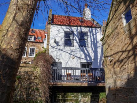 Back of a white house with red tiled roof and balcony with chairs and table surrounded by stone walls and trees at Roseberry Cottage in Robin Hood's Bay