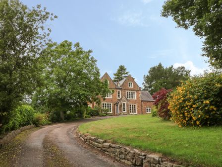 A driveway leading to a brick house surrounded by trees and bushes with flowers at The Old Vicarage in Tiverton