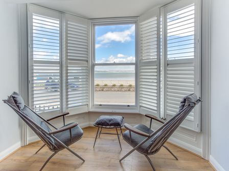 A sitting area with two chairs a footstool and plantation shutters by a bay window overlooking a waterfront at Low Tide Weymouth