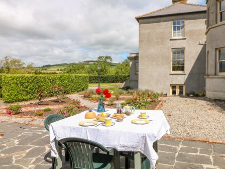An outdoor table with a white tablecloth set for tea and flowers in a vase in a garden beside a stone building at The Lodge at Raheengraney House in Raheengraney County Wicklow