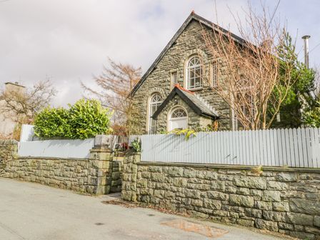 A stone house with arched windows behind a stone wall and white wooden fence with trees at Horeb Chapel House in Tanygrisiau