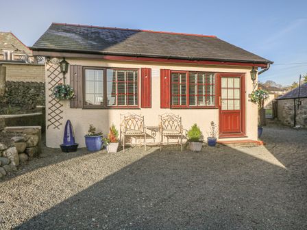 The exterior of a small building with two windows with shutters a door two chairs and several potted plants at Holly Cottage in Criccieth