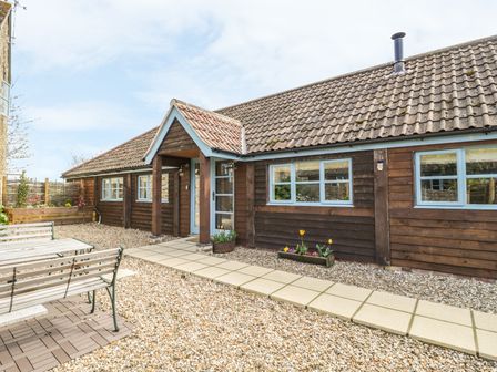 An outdoor area with a wooden bench and table set on gravel outside a brown wooden house with blue window frames and a tiled roof at Shippon Barn in Yeovil