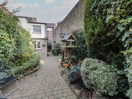 A paved garden pathway with bushes and potted plants leading to a white two-story house with windows and a door at Victoria Cottage in Amble