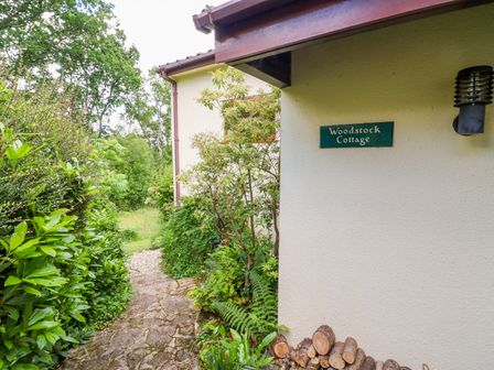 A stone path alongside greenery leading to a cream wall with a woodpile and a sign reading Woodstock Cottage at Birchill near Chardstock