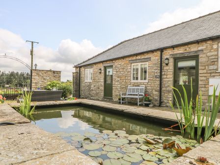 A stone building with two green doors and a bench next to a pond with lily pads at The Cottage in Thirsk
