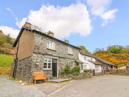 A stone and white cottage with a wooden bench outside surrounded by hills and trees at Tilberthwaite Farm Cottage in Coniston