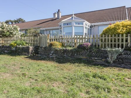 A house with a tiled roof and a conservatory behind a wooden picket fence and a stone wall with plants at Dune Haven in Goonhavern near Perranporth
