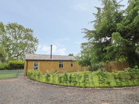A wooden building with windows and a chimney surrounded by grass trees and a gravel driveway at The Hen House in Stockwell Heath near Rugeley