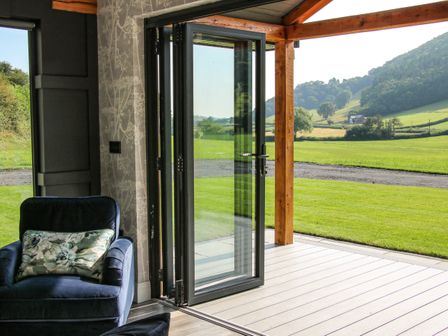 An armchair with a floral cushion next to a folding glass door opening to a patio and green fields at Meadow View in Meifod Powys