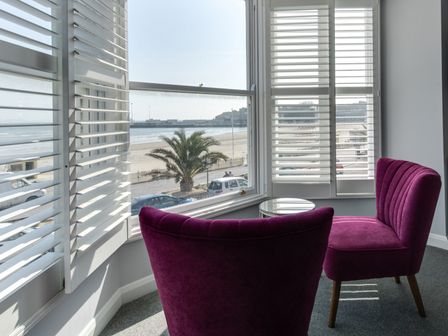 A corner with two purple chairs and a round glass table by windows with shutters overlooking the beach at High Tide in Weymouth