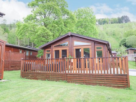 A wooden cabin with a fenced porch and trees behind at Fell View Lodge in Troutbeck