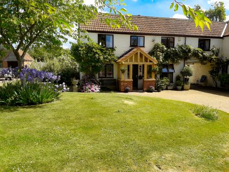 A garden with grass trees flowers and shrubs outside a house with windows and a wooden porch at Berrys Place Farm Cottage in Churcham