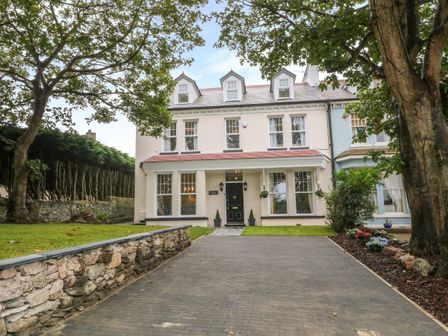 A front view of a three-story house with a driveway trees and flower beds at Lyngaran House in Holyhead