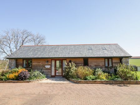 A single story wooden cottage with garden beds in front and a paved driveway at Kingcup Cottage in St Columb Major