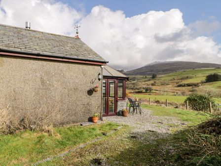 The exterior side of a stone house with a small patio table and chairs overlooking grassy hills at Melin Newydd in Porthmadog