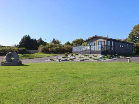 A wooden lodge with a glass balcony and outdoor seating on a paved area surrounded by grass and trees at Beech Lodge in Teigngrace near Newton Abbot