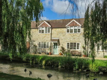 A stone house with a light blue door and white framed windows by a small river with ducks and overhanging trees at Duck Cottage in Sutton Poyntz