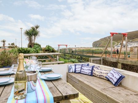 An outdoor dining area with a wooden table set with plates glasses a bottle and cushioned bench seating near a beach with swings at Offshore in Porthtowan
