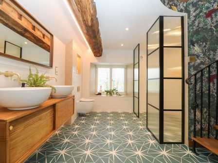 A bathroom with two white sinks on a wooden vanity a wall mirror a toilet a window with shutters patterned floor tiles and a shower with translucent panels at Tickmorend Farm in Nailsworth