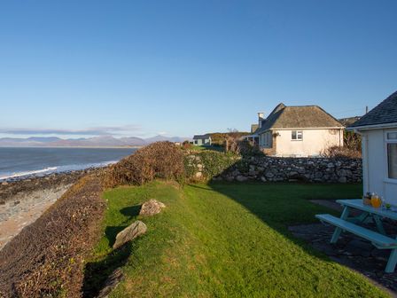 A grassy yard with a picnic table and houses near a rocky shoreline at Allt y Mor in Harlech