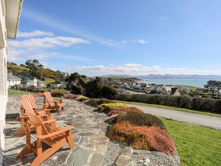 A stone patio with four wooden chairs overlooking houses hills and the sea at Bryn Deryn in Criccieth