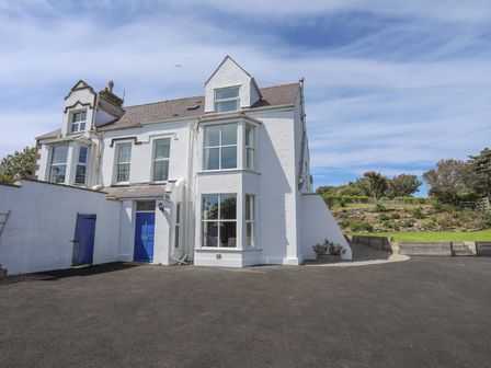 A white two-story house with large windows and a blue door with a paved driveway and garden area at Bryn Dale in Trearddur Bay