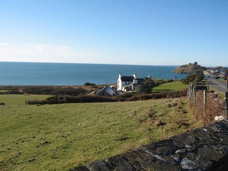 A coastal landscape with grass fields houses a stone wall and a road near Cae'r Dyni Caer Dyni in Criccieth