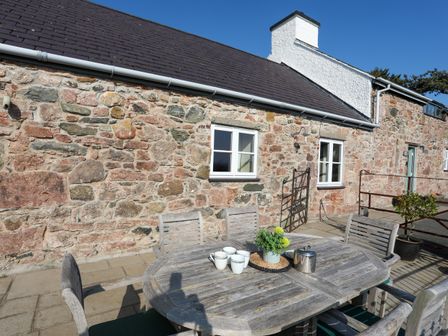 An outdoor patio with a wooden table and chairs with cups and a teapot against a stone cottage wall at Tan Twr - Chellow Cottage in Dwyran