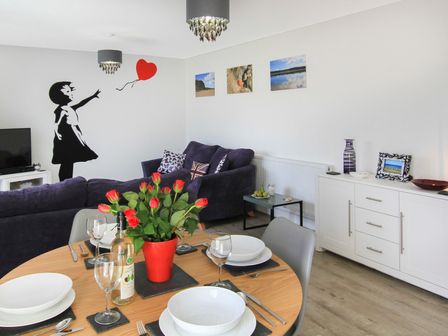 A dining area with a wooden table set with plates cups and cutlery and a vase of red flowers next to a living room with purple sofas wall art and a white sideboard at Cysgod Y Capel in Benllech