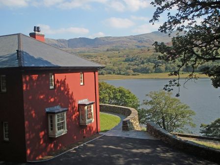 A red house next to a curved stone wall overlooking a lake and hills at Dinas Noddfa in Nant Gwynant