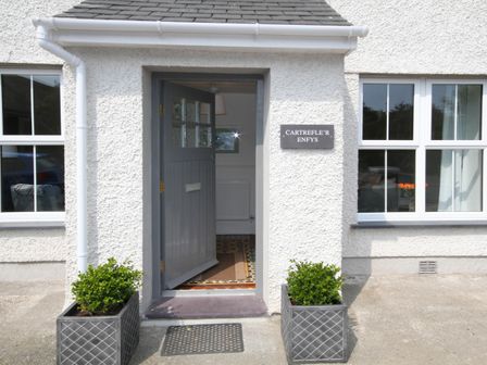 An open grey door at the entrance of a white building with two potted plants on either side at Cartrefle'r Enfys in Moelfre