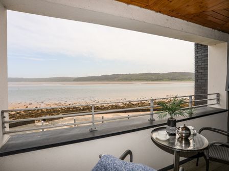 A balcony with a small table and two chairs overlooking a beach and hills at Four The Quay in Red Wharf Bay