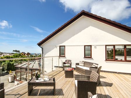 An outdoor deck with wicker furniture including chairs a sofa and tables with drinks on a tray and a house wall with windows at The Gables in Trearddur Bay