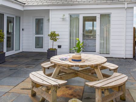A patio with a round wooden picnic table and benches a hat and plant on the table potted plants near white doors and windows at Hafod Llangoed in Llangoed