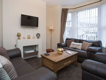 A living room with brown leather sofas a wooden coffee table a wall-mounted TV and a window with vertical blinds at Hirfron in Llanberis