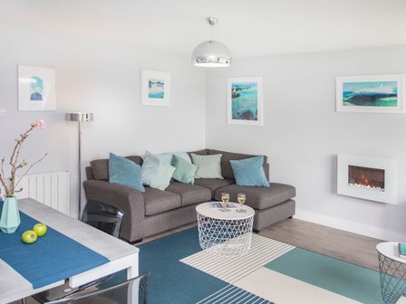 A living room with a gray sofa with blue and green pillows a white round wire coffee table with two glasses on it a wall-mounted fireplace and blue artwork at Isallt in Trearddur Bay