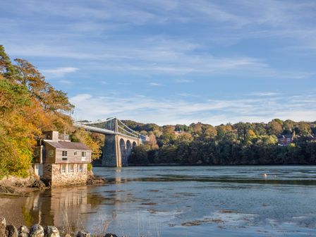 A house on a riverbank with a large stone bridge and trees across the water at Manadwyn in Menai Bridge