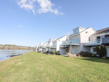 A row of waterfront houses with glass balconies facing a grassy area and water at Min Y Don in Y Felinheli