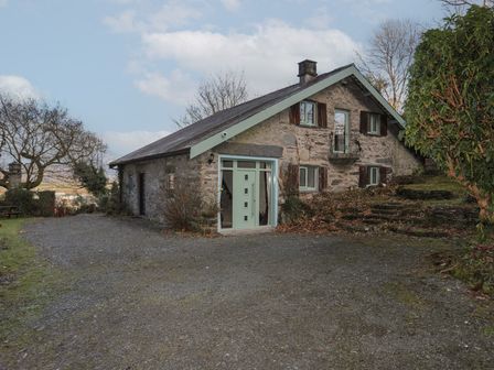 A stone cottage with light blue framed windows and door surrounded by trees and gravel at Morfa Lodge in Porthmadog