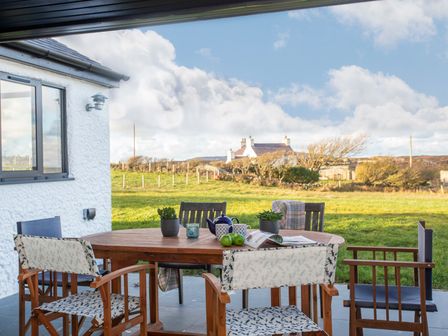 An outdoor patio with a wooden table set and chairs overlooking a grassy field and a white house in the distance at Mwythus in Rhoscolyn