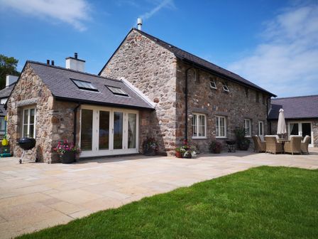 A stone house with patio doors and outdoor furniture on a paved area with grass in front at Penrhyn Barn in Dulas