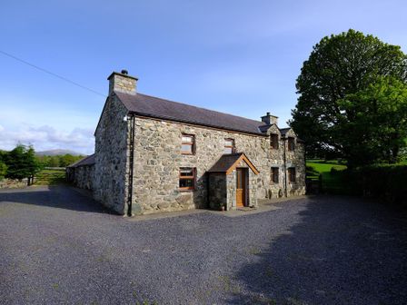 A stone cottage with a slate roof and wooden door surrounded by a gravel area and trees at Pen Y Bont in Pontllyfni
