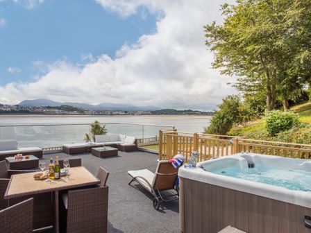 An outdoor patio with dining table and chairs a lounge area and a hot tub overlooking a body of water and distant buildings at The Mermaid in Brynsiencyn