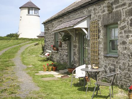 A stone cottage with green windows and a small outdoor table and chairs near a path with a tower in the background at Three Gates in Llangoed