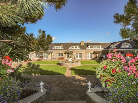 A stone building with multiple windows surrounding a courtyard with a central fountain and flowers at Tyn Towyn - Ty Gwyrdd in Trearddur Bay