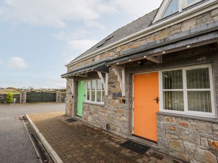 Stone exterior of a building with two doors one orange and one green with windows next to each door and a driveway in front at Tyn Towyn - Ty Oren in Trearddur Bay