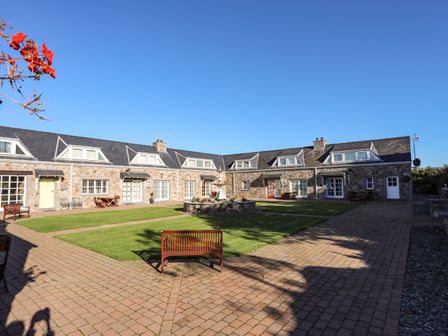 A courtyard with a stone fountain surrounded by benches and a stone building with multiple windows at Tyn Towyn - Ty Piws in Trearddur Bay
