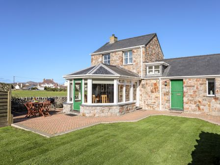 A stone house with green doors a paved patio with outdoor table and chairs and a lawn at Tyn Towyn - The Stables in Trearddur Bay